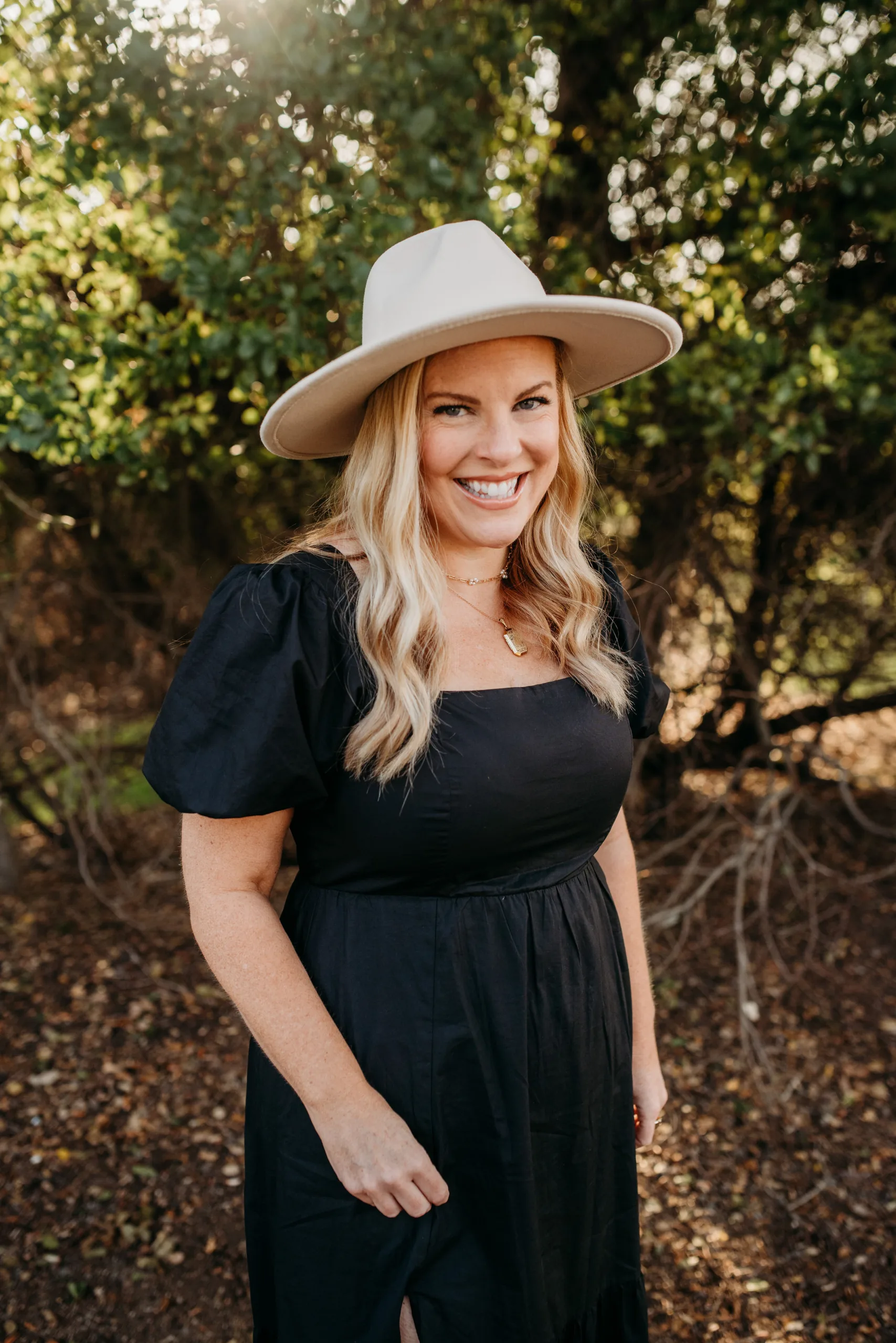 A woman in a black dress and white hat smiles in a sunlit wooded area.